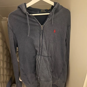 Blå hoodie från Ralph Lauren - Säljer en snygg blå hoodie från Ralph Lauren med dragkedja och huva. Den har en liten röd logga på bröstet och praktiska fickor framtill. Perfekt för en avslappnad stil.