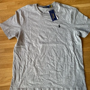 Grå t-shirt från Ralph Lauren - Säljer en stilren grå t-shirt från Ralph Lauren med den klassiska broderade loggan på bröstet. T-shirten har en normal passform och är tillverkad i mjuk bomull. Perfekt för en avslappnad look.