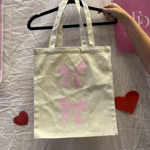 Beige shoppingväska med rosa rosetter - Vi är ett UF företag som säljer tygväskor med handmålade tryck på framsidan. Perfekt för att bära med sig det nödvändigaste med stil.