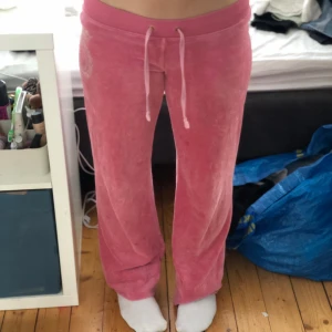 Rosa mjukisbyxor från Juicy Couture - Säljer ett par rosa mjukisbyxor från Juicy Couture med broderad logga och strassdetaljer på sidan. Byxorna har en bekväm passform med dragsko i midjan och är tillverkade i mjukt bomullsmaterial. Jag är 158 cm lång och har sytt upp byxorna så att de passar men om man är lite längre så kan man sprätta upp styngnen så blir byxorna 5cm längre 