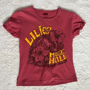 Röd t-shirt från Levi's med tryck - Säljer en cool röd t-shirt från Levi's med ett stort tryck på framsidan. Trycket har texten 'Lilies Music Hall' i gult och svart med blommotiv. Perfekt för en avslappnad stil. Passar S/M!