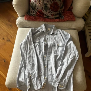 Grå skjorta från H&M - Säljer en stilren blå skjorta från H&M med normal passform. Skjortan har långa ärmar och knappar framtill samt på ärmsluten. Perfekt för en avslappnad men ändå snygg look.