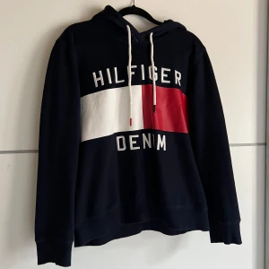Svart hoodie från Tommy Hilfiger - Säljer en svart hoodie från Tommy Hilfiger med det klassiska logotyptrycket i vitt och rött på framsidan. Tröjan har en justerbar huva med snören och långa ärmar. Perfekt för en avslappnad stil.
