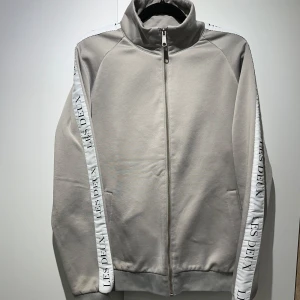 Grå kofta från Les Deux - Säljer en stilren grå jacka från Les Deux med dragkedja framtill. Jackan har långa ärmar med vita detaljer längs sidorna där märkesnamnet är tryckt. Perfekt för en casual look.