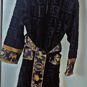 Svart morgonrock från Versace - Lyxig svart morgonrock från Versace med gulddetaljer och ett elegant mönster. Rocken har ett bälte i midjan och långa ärmar med dekorativa manschetter. Perfekt för en stilfull och bekväm hemmakväll.