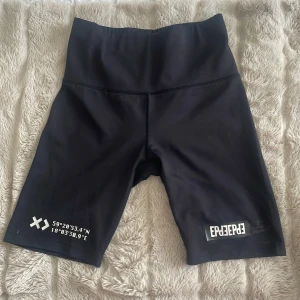 Svarta cykelshorts från Elly Pistol - Snygga svarta cykelshorts från Elly Pistol med hög midja och coola vita koordinatdetaljer. Perfekta för träning eller en avslappnad dag. Tillverkade i ett stretchigt syntetmaterial för bästa komfort.