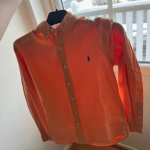 Orange skjorta från Ralph Lauren - Snygg orange skjorta från Ralph Lauren med klassisk krage och knappar framtill. Den har en broderad logga på bröstet och långa ärmar. Perfekt för en stilren look.