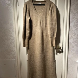 Beige långklänning - Elegant beige långklänning  i ett mjukt stickat material. Klänningen har en v-ringad hals och långa ärmar, perfekt passform. Den har en stilren design som passar till många tillfällen. Använd endast vid ett tillfälle.