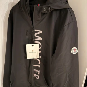  Moncler vindjacka  - Säljer en stilren svart jacka från Moncler med dragkedja och huva. Jackan har Monclers logotyp på ärmen och en stor text på framsidan. Den är i storlek XL men den sitter som en M/L