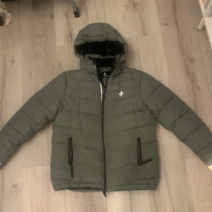 Grå dunjacka från Cross Sportswear - Säljer en grå dunjacka från Cross Sportswear. Jackan har en huva och dragkedja framtill samt två praktiska fickor med dragkedjor. Perfekt för kyliga dagar.