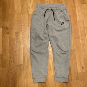 Grå nike tech byxor  - Snygga grå Nike tech byxor med svart logga på benet. Byxorna är storlek L för barn. Perfekta för en avslappnad stil. Byxorna har två normala fickor och en stor med dragkedja. Nypris 879kr. Säljer för att de inte passar längre. Kontakt vid minsta fundering