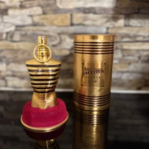 Jean Paul Gaultier Le Male Elixir - Elegant parfymflaska i form av en manlig torso med guld- och svartfärgade ränder. Flaskan står på en röd sammetsbas och kommer med en matchande guld- och svartfärgad cylinderförpackning. Perfekt för den som vill ha en lyxig och stilren doftupplevelse.