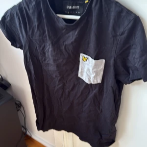 Svart t-shirt med ficka från Lyle & Scott - Snygg svart t-shirt från Lyle & Scott med en vit bröstficka och en gul broderad fjäril. Perfekt för en avslappnad stil. Kortärmad och tillverkad i mjukt material.