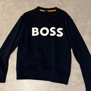 Mörk blå  sweatshirt från BOSS - Säljer en stilren svart sweatshirt från BOSS med deras logga i vitt på bröstet. Tröjan har långa ärmar och en klassisk rund halsringning. Perfekt för en avslappnad look.
