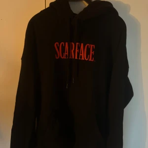 Svart Scarface hoodie - Säljer en svart hoodie med Scarface-tryck i rött på framsidan och en stor bild på baksidan. Perfekt för filmälskare som vill ha en cool och bekväm stil. Hoodien har en klassisk passform med långa ärmar och en justerbar huva.