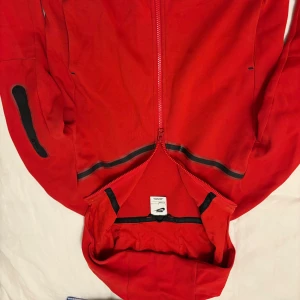 Röd nike tech  - Säljer en snygg röd jacka från Nike med dragkedja och svarta detaljer. Jackan har långa ärmar och en praktisk ficka med dragkedja på ärmen. Perfekt för en sportig look! Storlek se bild. 