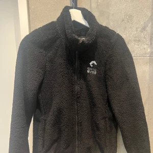 Svart  teddyjacka från Svea häst  - Mysig svart teddyjacka från Svea med dragkedja framtill. Perfekt för kyliga dagar med sin fluffiga och varma design. Jackan har en hög krage och långa ärmar, vilket gör den extra bekväm och skyddande mot vind.