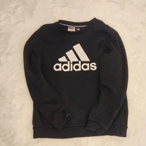 Svart sweatshirt från Adidas - Säljer en svart sweatshirt från Adidas med deras ikoniska logga i vitt på framsidan. Tröjan har långa ärmar och en klassisk rund halsringning. Perfekt för en sportig och avslappnad stil.
