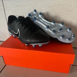 Nike Legend 10 Academy FG/MG fotbollsskor EU 38,5 - Snygga svarta Nike Legend 10 Academy FG/MG fotbollsskor med vit och blå detaljering. Skorna har en klassisk snörning och är designade för både fasta och multi-ground underlag. Nyskick! Storlek 38,5