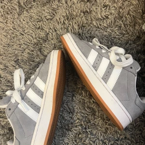 Grå Adidas sneakers - Snygga grå sneakers från Adidas med vita detaljer och klassiska tre ränder på sidan. Skorna har en gummisula i brun nyans och snörning framtill. Perfekta för en avslappnad stil. Knappt använda. 