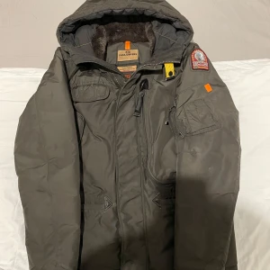 Grön parkajacka från Parajumpers - Säljer en grön parkajacka från Parajumpers med huva och flera praktiska fickor. Jackan har en broderad patch på ärmen och dragkedja framtill. Perfekt för kalla dagar.