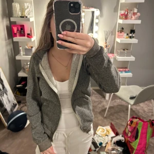 Grå hoodie med dragkedja - Säljer en mysig grå hoodie med dragkedja framtill. Perfekt för en avslappnad stil och enkel att matcha med andra plagg. Den har långa ärmar och en bekväm passform.