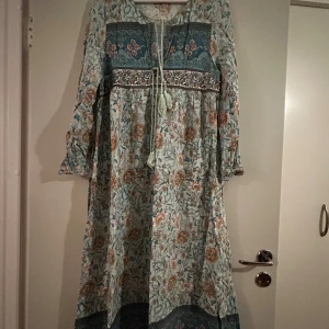 Blommig långklänning med knytband - Charmig långklänning med blommigt mönster i blå och beige toner. Klänningen har långa ärmar och ett knytband framtill som ger en bohemisk känsla. Perfekt för en avslappnad stil.