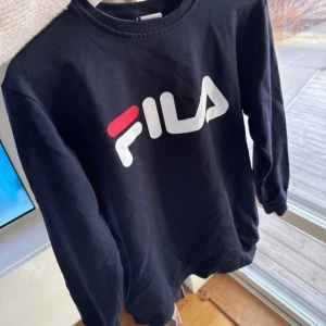 Svart sweatshirt från Fila - Snygg svart sweatshirt från Fila med deras ikoniska logga i vitt och rött på bröstet. Tröjan har långa ärmar och en rund halsringning. Perfekt för en avslappnad stil.