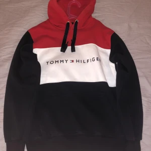 Flerfärgad hoodie från Tommy Hilfiger - Säljer en snygg flerfärgad hoodie från Tommy Hilfiger. Den har en röd, vit och svart design med märkets logga på bröstet. Hoodien har en justerbar huva med svarta snören och en praktisk magficka. Perfekt för en avslappnad stil.