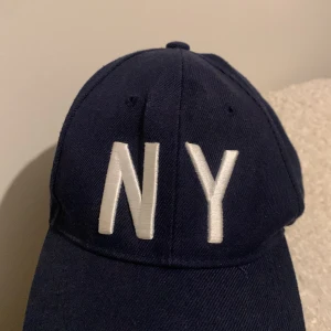 Mörkblå keps med NY-brodyr - Snygg mörkblå keps med vit brodyr där det står NY framtill. Klassisk böjd skärm och justerbar snapback baktill för perfekt passform. Tillverkad i bomullstyg som känns både bekvämt och stilrent. Perfekt accessoar för en sportig och urban look.