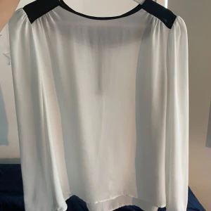 Vit blus med svarta detaljer och dragkedja - Säljer en stilren vit blus med svarta detaljer vid axlar, halsringning och ärmslut. Blusen har lång ärm och en snygg guldig dragkedja bak. Tunt, lätt material som ger en luftig känsla. Perfekt till jeans eller kjol för en clean look.