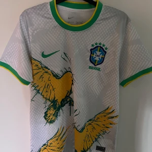 Vit Brasilien fotbollströja Nike - Vit Brasilien fotbollströja från Nike med gröna och gula detaljer på krage och ärmslut. Framsidan har ett stort gult fågelmotiv och CBF-emblem på bröstet. Tillverkad i lätt och ventilerande material, perfekt för match eller träning.