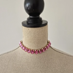 Chokerhalsband med rosa stenar - Statement chokerhalsband med stora, rektangulära rosa stenar infattade i guldfärgad metall. Kedjan är tunn och elegant, vilket gör att de glittrande stenarna verkligen står ut. Perfekt för dig som vill sticka ut med färg och bling.