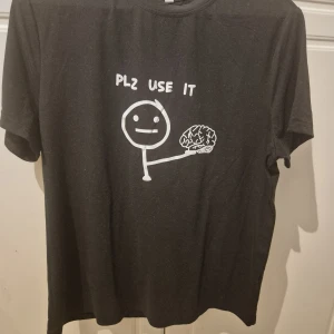 Svart t-shirt med meme-tryck - Svart t-shirt med ett vitt meme-tryck framtill som föreställer en streckgubbe som håller en hjärna och texten 'PLZ USE IT'. Klassisk rund hals och korta ärmar. Perfekt för dig som gillar humor och vill sticka ut.