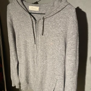 Grå hoodie från Saylus med dragkedja - Mysig grå hoodie från Saylus i stickat material. Tröjan har huva med snörning, dragkedja framtill och två fickor. Perfekt för dig som gillar en avslappnad och enkel stil.