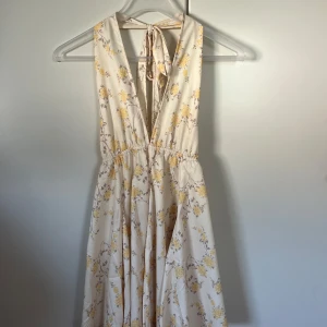 Blommig långklänning med knyt i ryggen - Superfin långklänning i ljusbeige med gula blommor och v-ringning. Klänningen har omlottkänsla, knyt i nacken, flowig kjol och öppen rygg med extra knytband. Bara använd en gång.