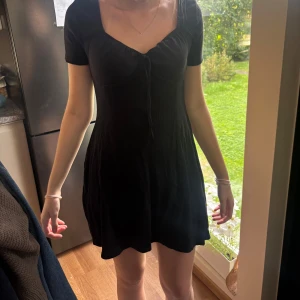 Svart kortklänning med snörning - Supersöt svart kortklänning med snörning framtill och v-ringad hals. Klänningen har korta ärmar och en lätt utställd kjol som ger en skön och avslappnad vibe. Perfekt för dig som gillar enkel men snygg stil.