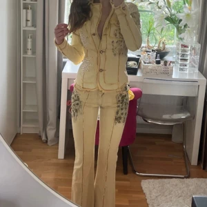 Unikt jeansset  - Unikt set från Daisy's i ljusgult. Både toppen och jeansen har broderade detaljer, pärlor och fransar längs sömmarna samt dekorativa mönster i svart och vitt. Materialet är bomull och denim, och passformen är normal. Jeansen är Bootcut med låg midja.