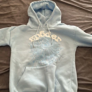 Ljusblå hoodie från Sp5der - Säljer en ljusblå hoodie från Sp5der med vit logga och ett stort spindelnätsmönster på bröstet. Tröjan har huva med snörning, magficka och ribbade muddar. Perfekt för dig som gillar streetwear och vill sticka ut lite extra. Står storlek m men det passar till storlek s priset kan diskuteras 