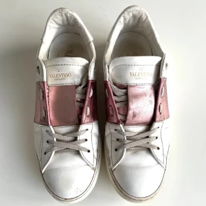 Valentino Garavani sneakers med rosa detalj - Snygga vita sneakers från Valentino Garavani med metallicrosa band över snörningen. Skorna har rund tå, platt sula och är tillverkade i skinn. Perfekta för dig som vill sticka ut med en lyxig touch och cool design.