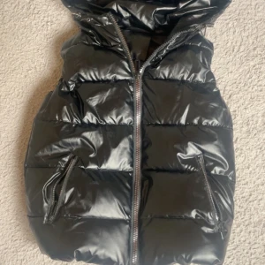Svart glansig dunväst med huva - Snygg svart dunväst med glansig finish och stor huva. Västen har två fickor med dragkedja och en rejäl dragkedja framtill. Perfekt för lager-på-lager och streetstyle.