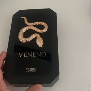Veneno parfym från French Avenue - Lyxig parfym i svart, kantig flaska med en detaljerad guldfärgad orm som dekoration. Kommer i en exklusiv svart ask med gulddetaljer och samma orm-motiv. Flaskan har svart lock och genomskinliga sidor som visar den gyllene vätskan inuti.