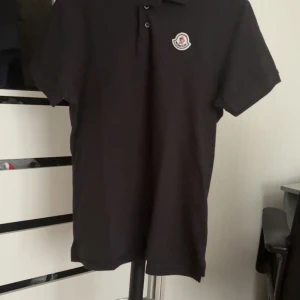 Mocler piké - Svart pikétröja från Moncler med klassisk krage och två knappar framtill. T-shirten har korta ärmar och en broderad Moncler-logga på bröstet. Tillverkad i mjuk bomull för en clean och stilren look. PRIS KAN DISKUTERAS 