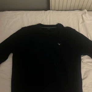 Svart stickad tröja från Gant - Klassisk svart stickad tröja från Gant med rund hals och diskret broderad logga på bröstet. Tröjan har lång ärm och en clean look som funkar till allt. Perfekt för dig som gillar stilrena och tidlösa plagg.