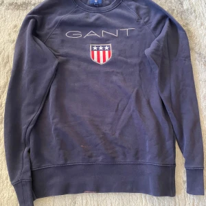 Mörkblå sweatshirt från GANT - Mörkblå sweatshirt från GANT med broderad logga och sköld med stjärnor och ränder på bröstet. Klassisk rund halsringning och långa ärmar.               Ord pris: 550kr 