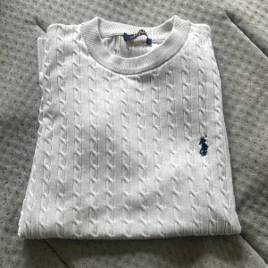 Vit kabelstickad tröja Polo Ralph Lauren - Vit kabelstickad tröja från Polo Ralph Lauren med korta ärmar och rund hals. Klassisk design med broderad marinblå logga på bröstet. Tröjan är i bomull och har ribbade muddar vid hals, ärmslut och nederkant. Perfekt för en clean och stilren look. Passar även M 