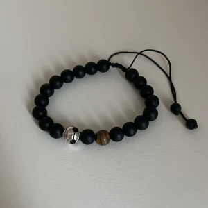 Svart pärlarmband med fotbollspärla - Helt ny! Coolt armband med svarta matta pärlor, en brun pärla och en silverfärgad pärla formad som en fotboll. Justerbar snörning för perfekt passform. Perfekt accessoar för dig som älskar sportig stil och detaljer som sticker ut.