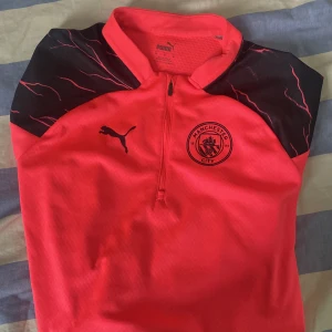 Manchester City tränings-topp Puma M - Cool Manchester City tränings-topp från Puma i stark röd färg med svarta detaljer och klubbmärke på bröstet. Halvlång dragkedja framtill, svarta axelpartier med mönster och tillverkad i lätt, ventilerande polyester. Perfekt för fotbollsträning eller support.