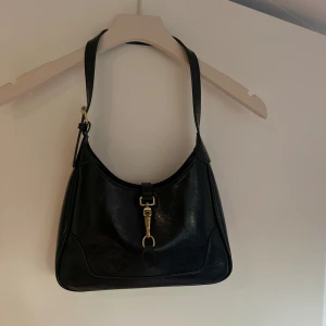 Svart axelväska i skinn med gulddetalj - Snygg svart axelväska i skinn med stilren form och guldfärgad metallspänne framtill. Väskan har en justerbar axelrem och minimalistisk design som passar till många outfits. Perfekt storlek för det viktigaste och ger en classy vibe.