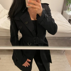 Svart trenchcoat från Burberry S (äkta) - Snygg svart trenchcoat från Burberry med klassiskt rutigt foder. Jackan har bälte i midjan, dubbelknäppning och detaljer vid ärmsluten. 